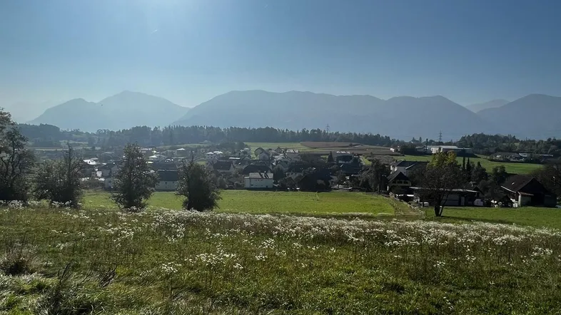 Sonnenbeschienene Landschaft mit weiten Feldern, einem Dorf im Tal und majestätischen Bergen im Hintergrund.