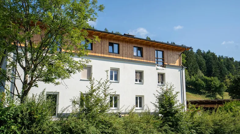 Modernes Gebäude mit weißer Fassade und Holzverkleidung unter blauem Himmel, umgeben von Bäumen.