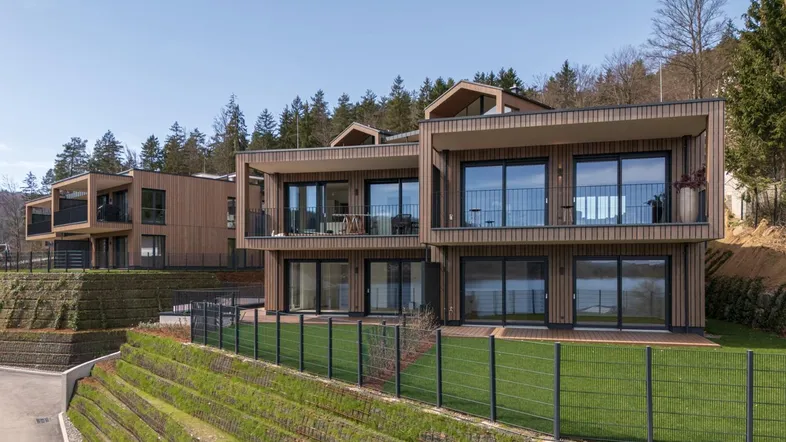 Moderne Wohngebäude mit Holzfassaden, großen Fenstern und Balkonen, eingebettet in eine grüne Hanglage mit Seeblick.