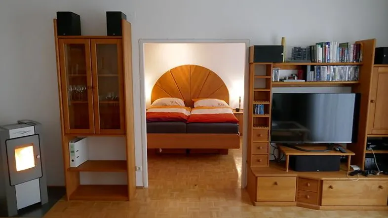 Wohnbereich mit Kaminofen und Blick in das angrenzende Schlafzimmer, ausgestattet mit Holzmöbeln.