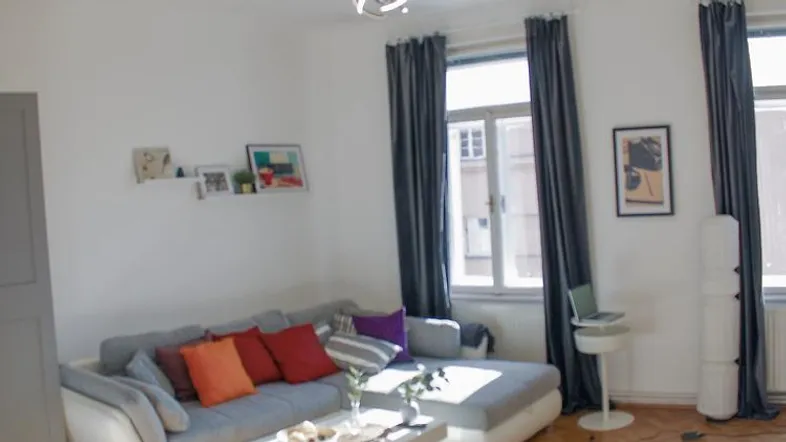 Helles Wohnzimmer mit gemütlichem L-Sofa, bunten Kissen und Holzboden, durch große Fenster gut belichtet.