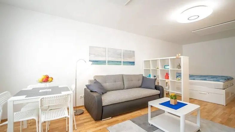 Helles Wohnzimmer mit Sofa, Esstisch und Regal, Teil eines Studio-Apartments mit Schlafbereich.