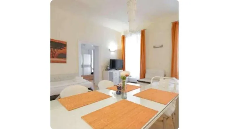 Helles Zimmer mit Esstisch und Wohnbereich, orangefarbene Vorhänge sorgen für Farbakzente.