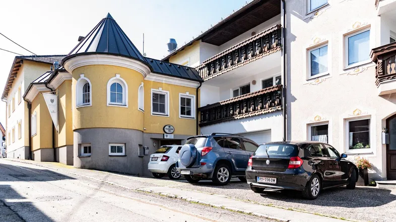 Traditionelles Gebäude mit gelber Fassade, Turm und Parkplätzen an einer leicht ansteigenden Straße.