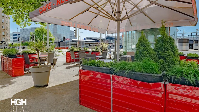 Großer Außenbereich eines Cafés mit Tischen, Stühlen, Sonnenschirm und Pflanzen in roten Blumenkästen.