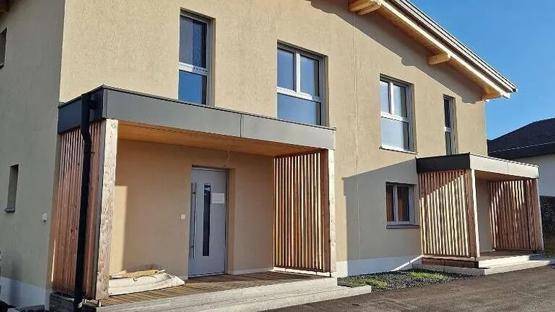 Modernes Doppelhaus mit heller Fassade, Holzdetails und überdachtem Eingangsbereich unter blauem Himmel.