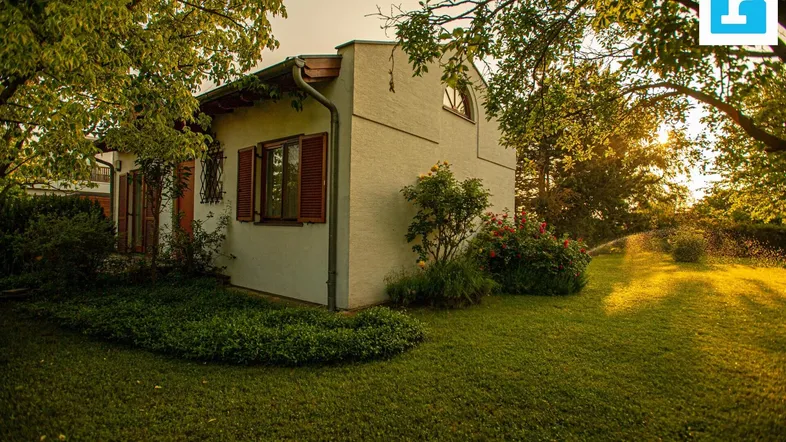 Kleines Haus umgeben von einem gepflegten Garten mit Bäumen und Sträuchern im warmen Abendlicht.