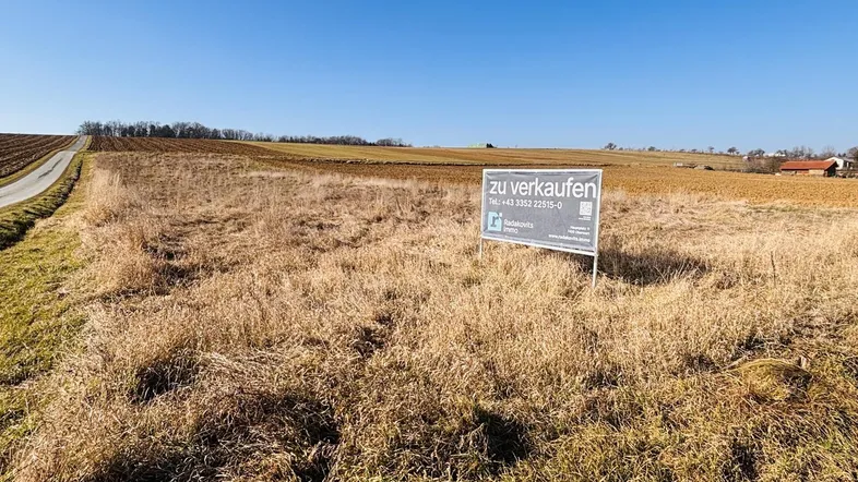 Ein 'Zu Verkaufen'-Schild steht auf einem trockenen Feld, umgeben von ländlicher Landschaft unter klarem blauem Himmel.