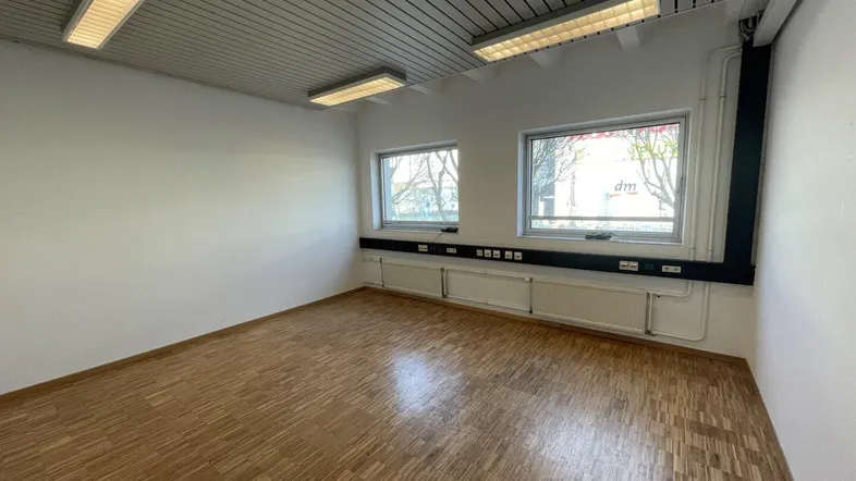 Heller, leerer Büroraum mit Parkettboden, zwei Fenstern und Deckenleuchten, ideal für gewerbliche Nutzung.