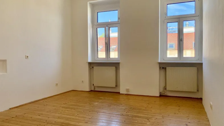 Heller Raum mit Holzdielenboden und zwei großen Fenstern, die viel Tageslicht hereinlassen.
