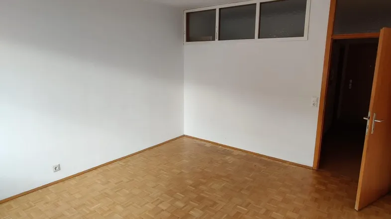 Helles, leeres Zimmer mit Parkettboden und einer Tür, die in einen weiteren Raum führt.