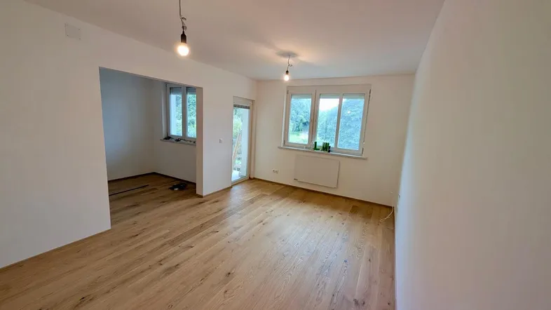 Helles Wohnzimmer mit neuem Holzboden und Zugang zu einem weiteren Raum sowie einem Fenster mit Blick ins Grüne.