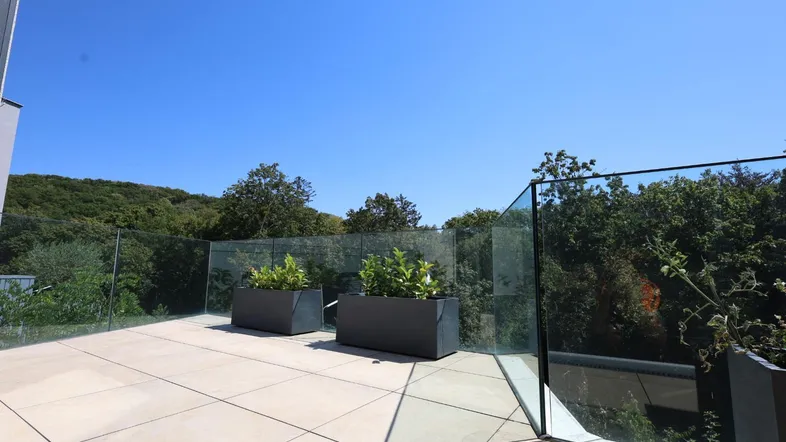 Große, moderne Terrasse mit Glasgeländer, Pflanzenkübeln und weitem Blick ins Grüne.