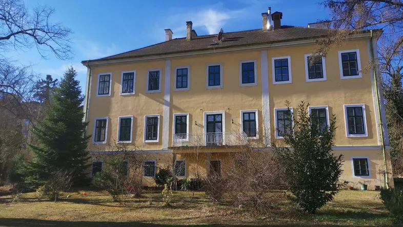 Historisches Gebäude mit gelber Fassade, umgeben von Bäumen und einem weitläufigen Garten.