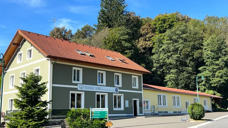 Traditionelles Gebäude mit grüner Fassade und rotem Ziegeldach, umgeben von Bäumen und einem Parkplatz.
