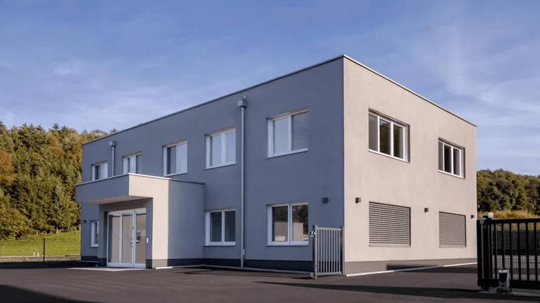 Modernes Bürogebäude mit grauer Fassade und großen Fenstern, umgeben von Natur unter blauem Himmel.