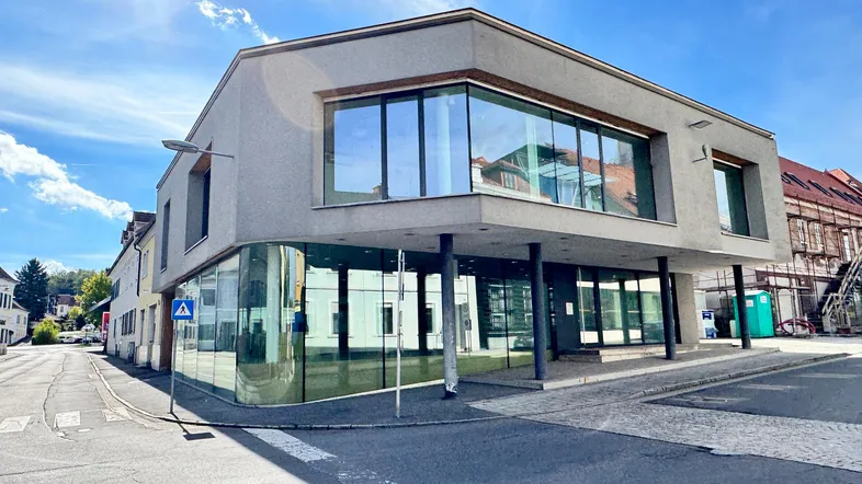 Modernes Geschäftsgebäude mit großen Glasflächen an einer Straßenecke unter blauem Himmel und Sonnenschein.