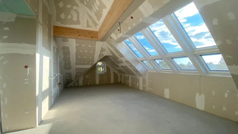 Heller Dachbodenraum im Rohbau mit sichtbaren Holzbalken und mehreren Dachfenstern, bereit für den Ausbau.