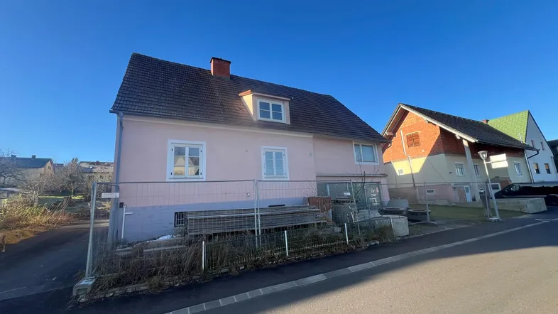 Rosa verputztes Haus mit Satteldach und weißen Fenstern, umgeben von einem Bauzaun.