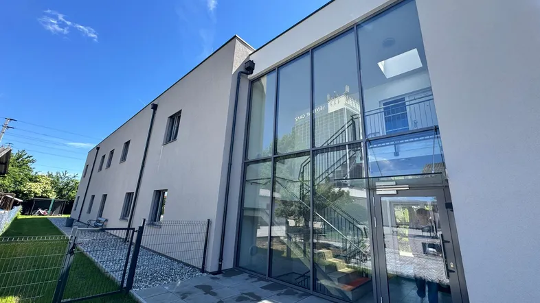 Moderne Gebäudefassade mit großer Glasfront und Eingangsbereich, umgeben von Grünflächen und einem Zaun.