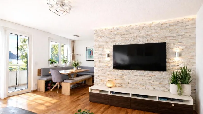 Helles Wohn- und Esszimmer mit Holzboden, Steinwand und Zugang zum Balkon.