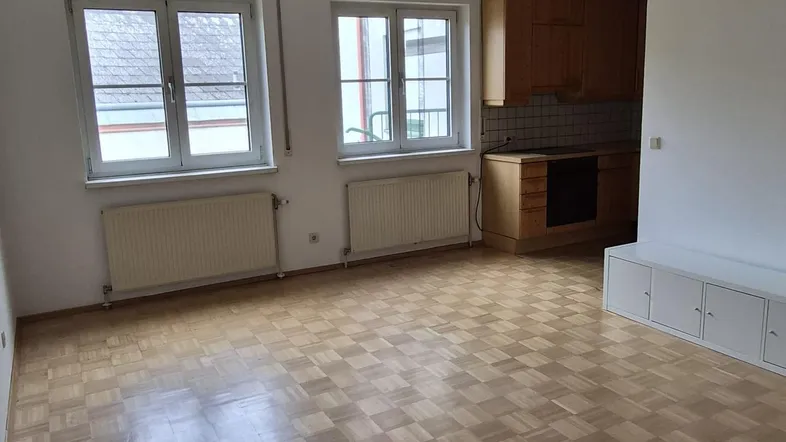 Heller Wohn- und Essbereich mit Parkettboden, zwei Fenstern und angrenzender Küche.