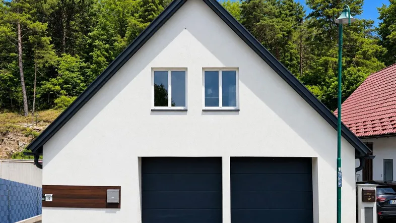 Moderne Hausfassade mit zwei Garagentoren und Satteldach, umgeben von Bäumen.
