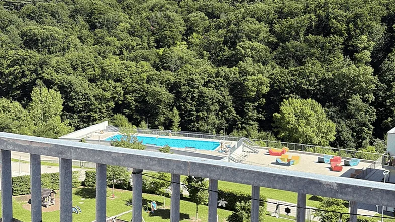 Panoramablick vom Balkon auf einen Gemeinschaftspool, Spielplatz und grüne bewaldete Berge unter blauem Himmel.