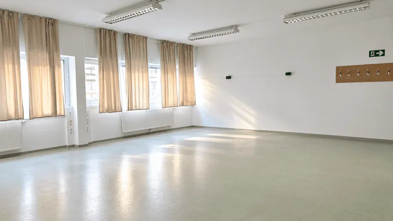 Heller, leerer Raum mit mehreren Fenstern, Vorhängen und einer Garderobenleiste an der Wand.
