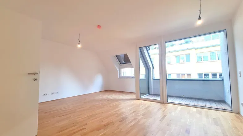Helles, leeres Zimmer mit Parkettboden, Dachschrägen und Zugang zu einem kleinen Balkon.