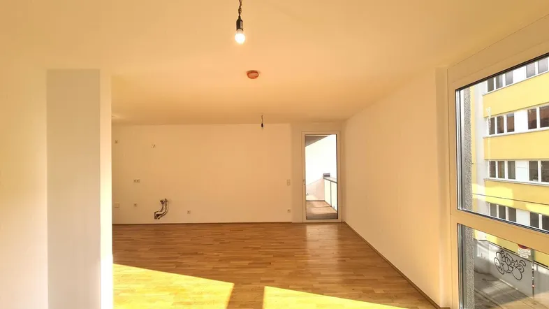 Helles, leeres Wohnzimmer mit Parkettboden und großem Fenster, das viel Sonnenlicht hereinlässt.