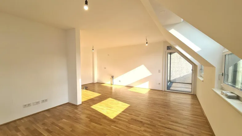 Helles, leeres Zimmer mit Holzparkettboden und Zugang zu einem Balkon, durchflutet von Sonnenlicht.