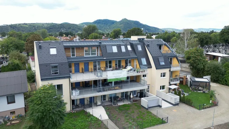 Luftaufnahme eines modernen Mehrfamilienhauses mit mehreren Balkonen und Dachschrägen in grüner Umgebung.