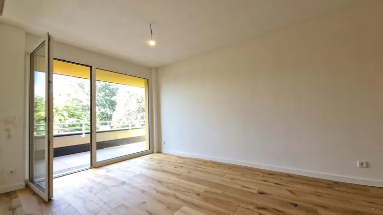 Helles Zimmer mit Holzboden und Zugang zu einem sonnigen Balkon, ideal für Wohn- oder Schlafbereich.