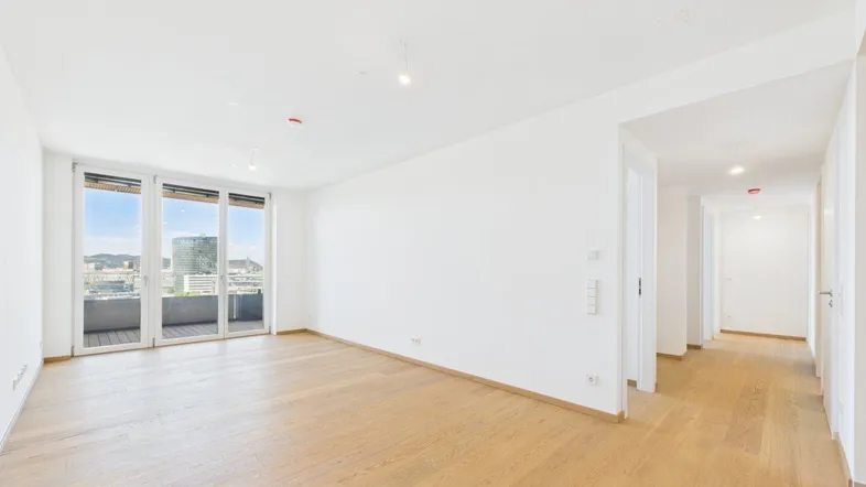 Helles, leeres Wohnzimmer mit Parkettboden und Zugang zu einem Balkon mit Stadtblick.