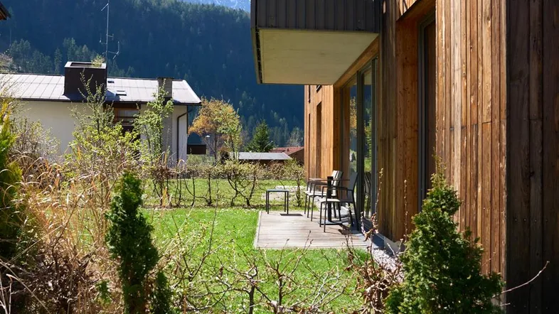 Moderne Holzfassade mit kleiner Terrasse, umgeben von Grünflächen und mit Blick auf die Berge.