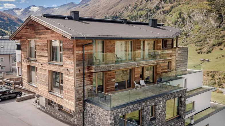 Modernes Chalet-Gebäude mit Holzfassade, Steinsockel und Glasbalkonen vor alpiner Bergkulisse.