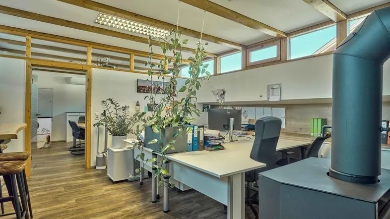 Helles und modernes Büro mit Holzbalken, großen Fenstern und einem Kaminofen als Blickfang.