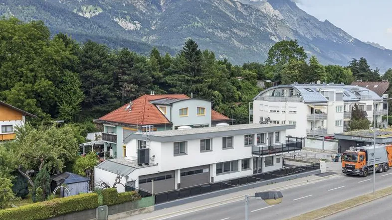 Luftaufnahme des modernen Gebäudes an einer Straße, umgeben von Bäumen und einer beeindruckenden Bergkulisse.