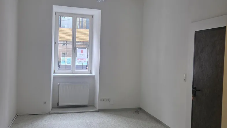 Heller, leerer Raum mit einem Fenster, das Tageslicht hereinlässt, und einem Heizkörper darunter.