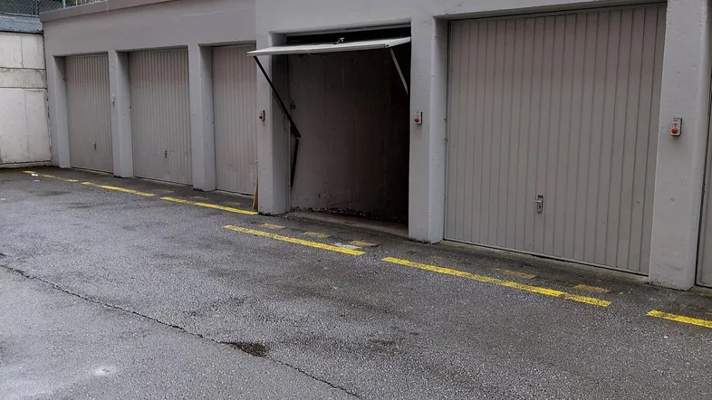 Reihe von Garagenboxen mit einer offenen Tür und asphaltiertem Vorplatz vor einem Wohngebäude.