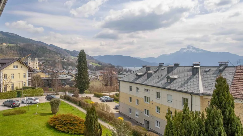 Panoramablick auf die umliegende Berglandschaft, einen See und Wohngebäude unter bewölktem Himmel.