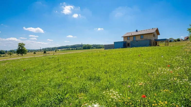 Ein im Bau befindliches Haus in einer weiten grünen Landschaft unter blauem Himmel mit blühenden Wiesen.