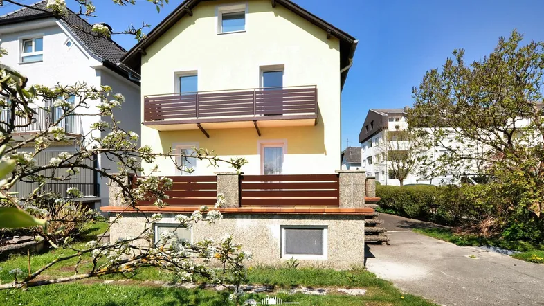 Gelbes Einfamilienhaus mit Balkon und kleinem Vorgarten unter blauem Himmel.
