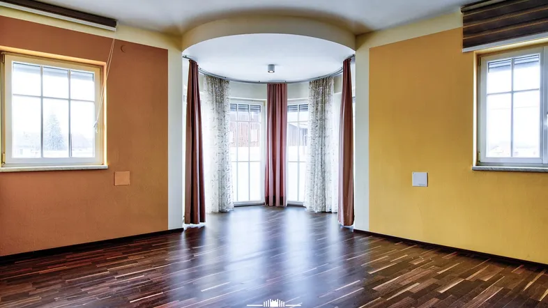 Geräumiges Wohnzimmer mit dunklem Parkettboden, farbigen Wänden und einem hellen Erkerbereich.