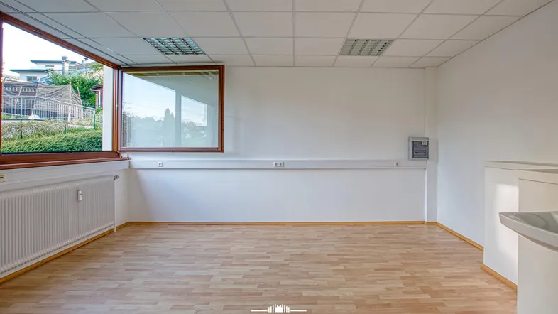 Heller Büroraum mit Laminatboden, großen Fenstern und einem Ausblick ins Grüne.