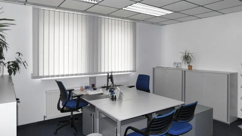 Helles Büro mit mehreren Arbeitsplätzen, vertikalen Jalousien und abgehängter Decke.