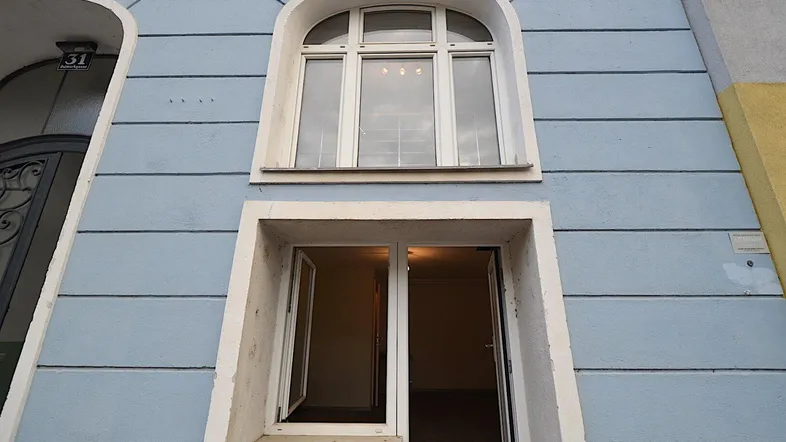 Blaue Fassade eines Altbaus mit einem großen Bogenfenster und einer offenen Eingangstür zum Innenbereich.