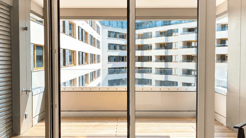 Helles Zimmer mit Holzboden und Zugang zu einem großen Balkon mit Blick auf moderne Gebäude.