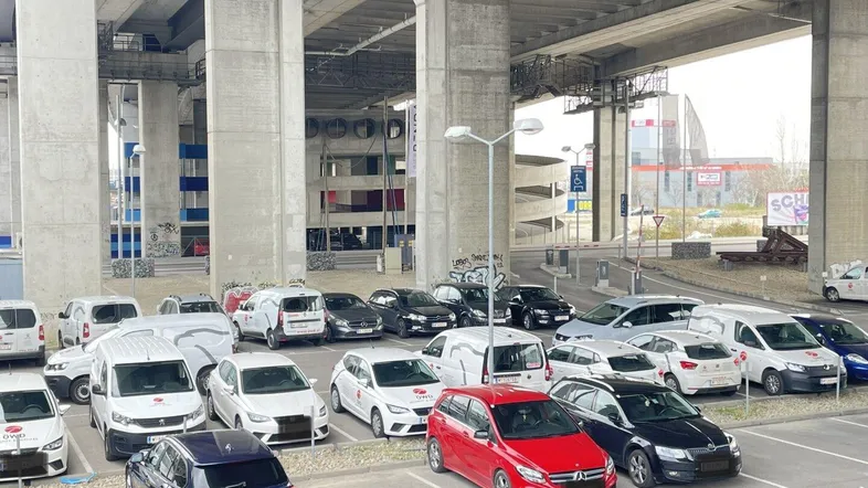 Großer Außenparkplatz unter einer massiven Betonbrückenkonstruktion mit zahlreichen abgestellten Fahrzeugen in urbaner Umgebung.